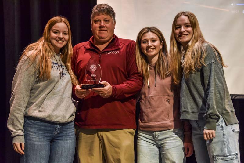 Lyndon Institute Director of Athletics Eric Berry (second from left) is photographed here with current varsity girls basketball team captains Ary Parker (far left), Brooke'lyn Robinson (second from right), and Molly Smith (far right) at an all-school assembly on the LI campus honoring coach Berry's 200 wins as head coach of LI's varsity girls basketball team, and his 300 all-time career wins.